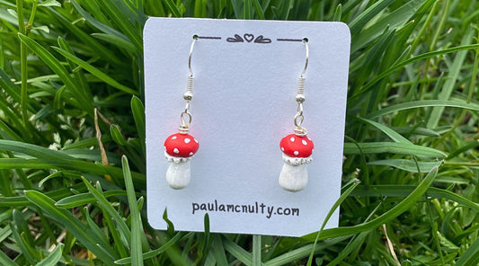 Mini red capped mushroom earrings on an earring card nestled in grass.