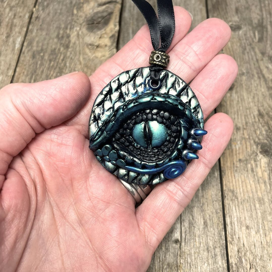 A round dragon eye ornament with metallic green gold and blue coloring with black satin ribbon held in hand