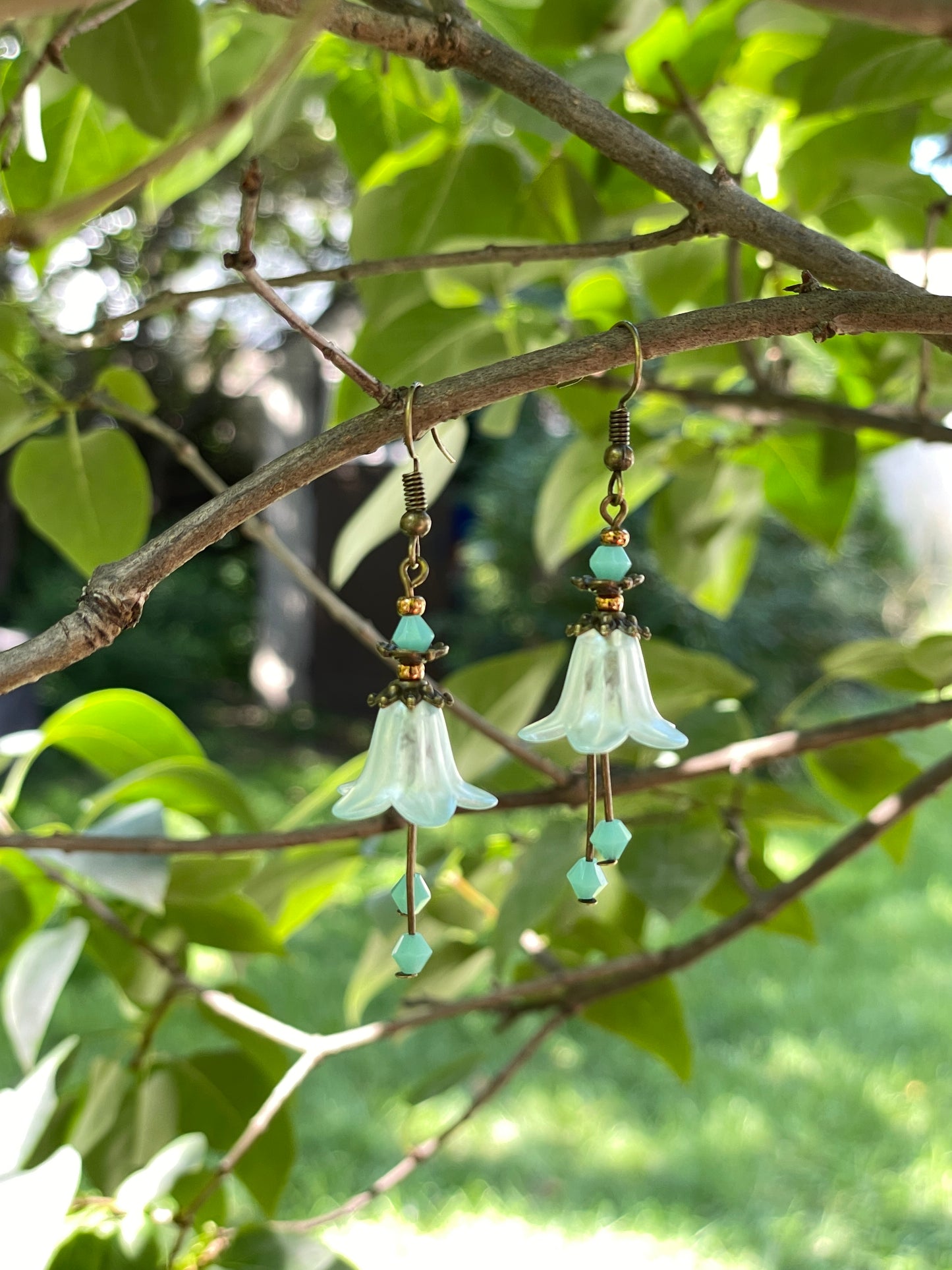 light green bell flower earrings with green crystals and antique bronze fitinish. Earrings are hanging on a tree branch.