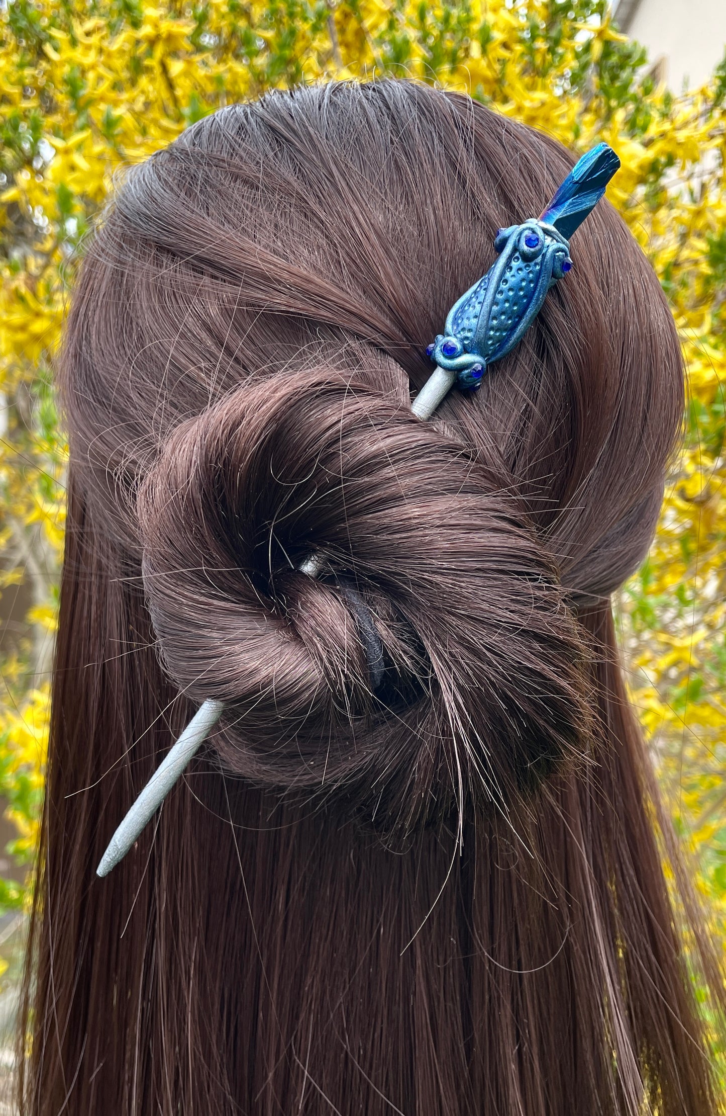 Model wearing blue raw crystal hair stick with hand painted clay details and crystals resting on a pebble with a black flower to the side.
