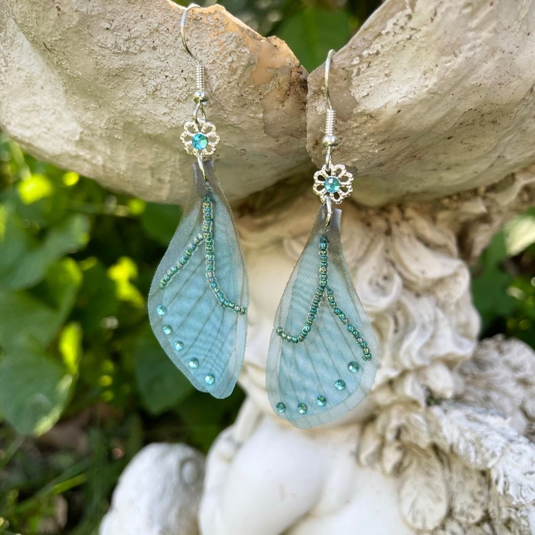 Blue chiffon butterfly wing earrings with silver crystal flower, charm and crystals on the wings hanging from a garden ornament.