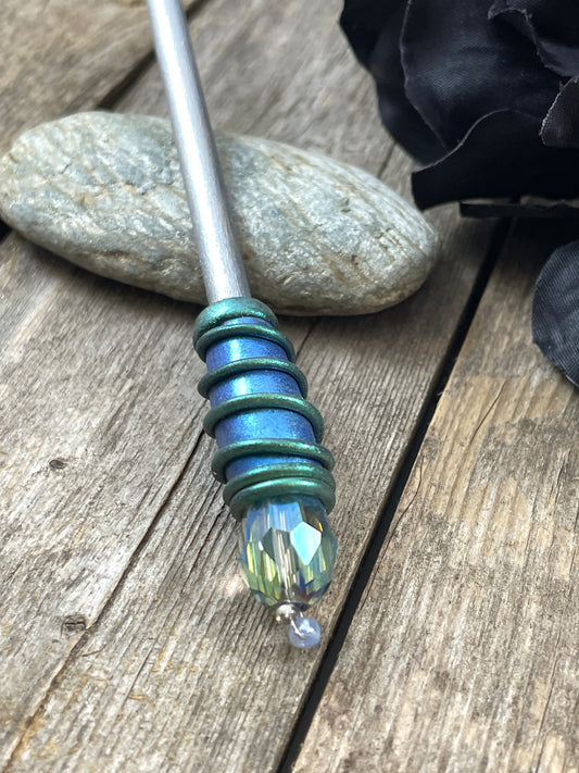 Hair stick with blue and green spiral clay hand painted details with green AB crystal. Resting on a pebble with a black flower to one side.