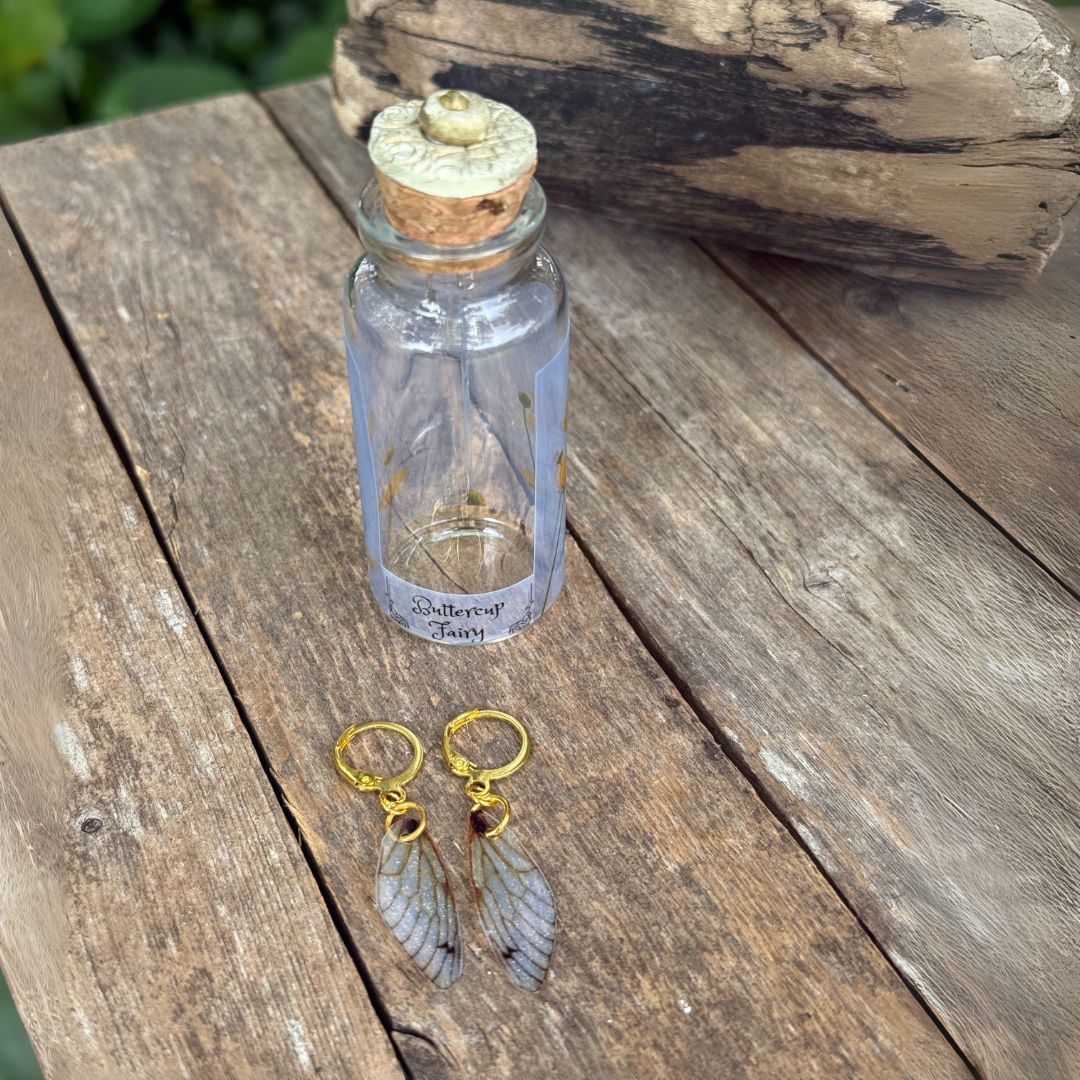 Small glass bottle with cork lid on a wooden surface, accompanied by gold and gray earrings.
