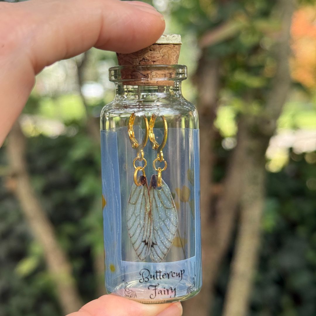 Glass bottle with cork lid containing butterfly-themed earrings, held by a hand with a blurred green outdoor background.