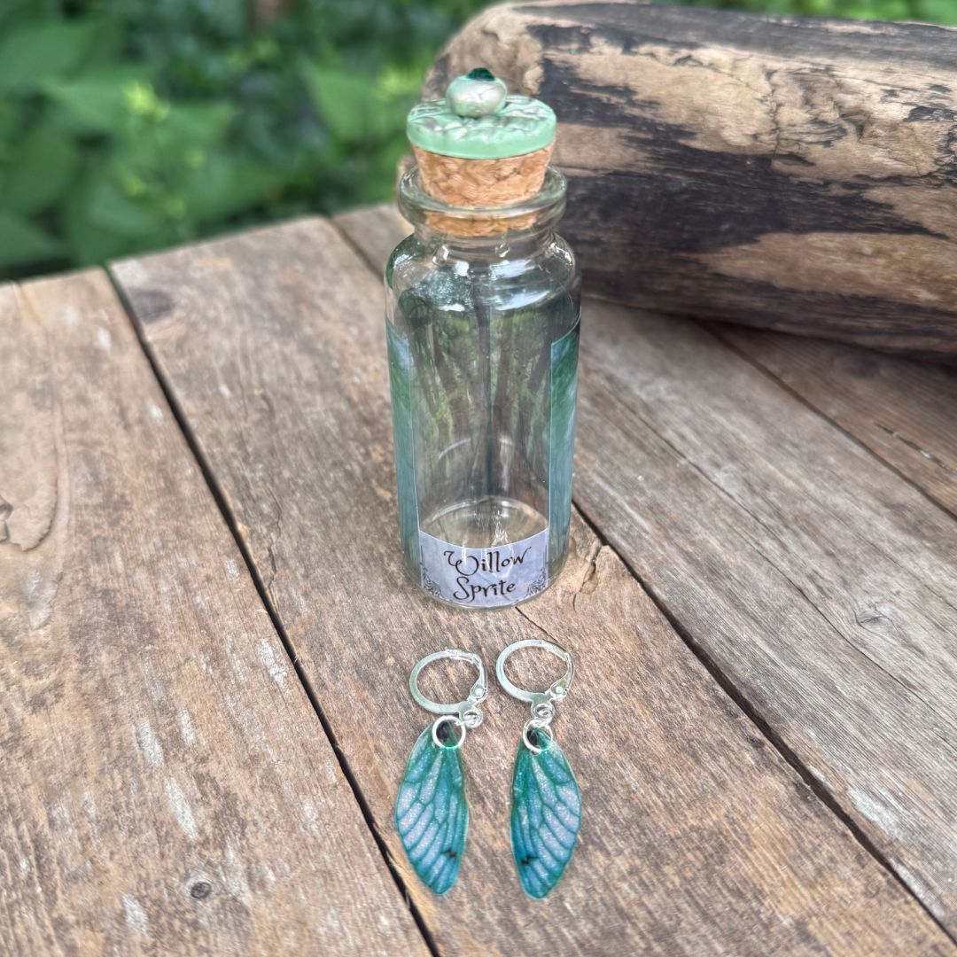 Small glass bottle labeled 'Willow Sprite' with cork lid on a wooden surface, accompanied by green wing-shaped earrings.
