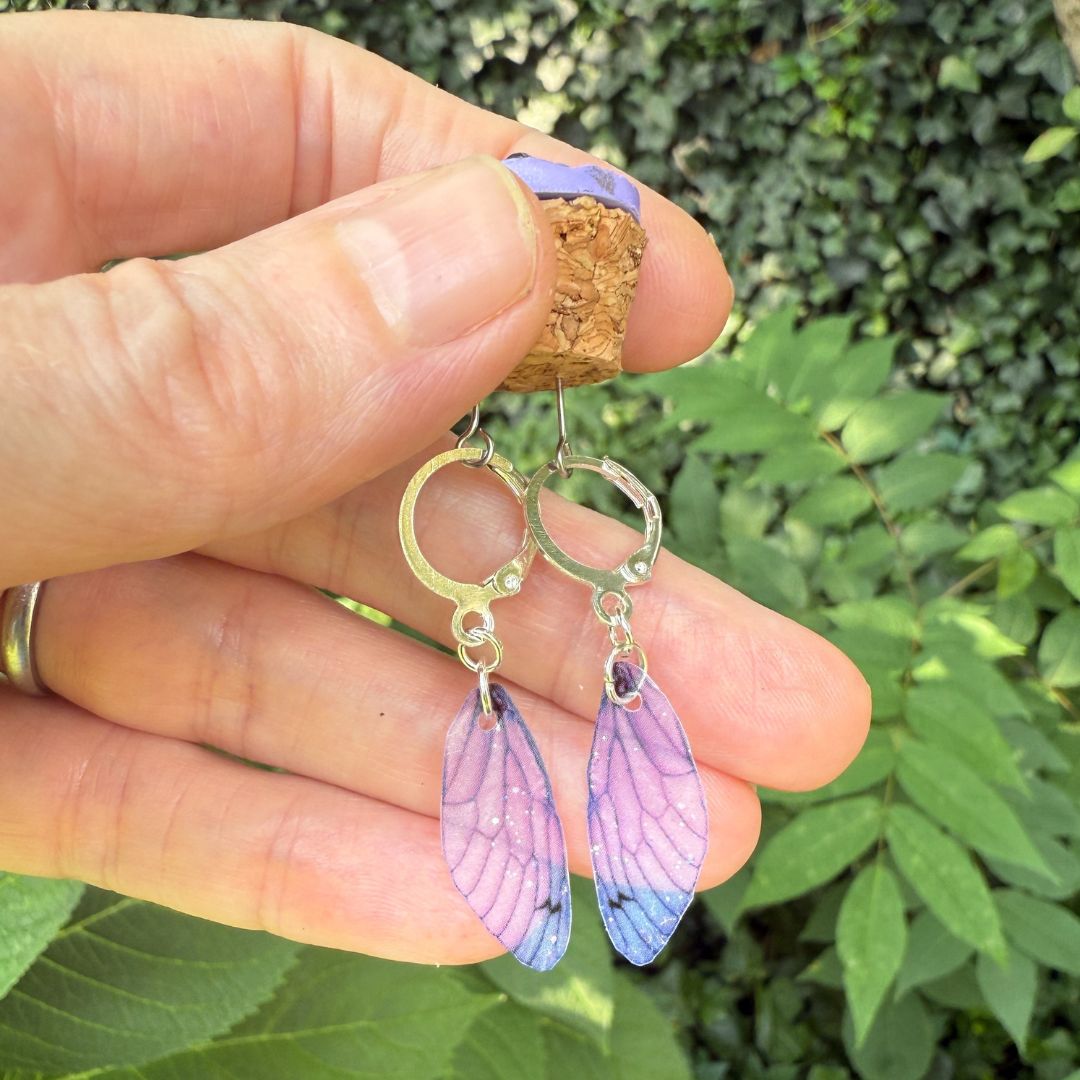 Hand holding a pair of purple wing-shaped earrings with a green leafy background