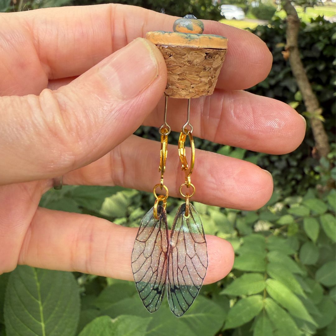 Hand holding a pair of earrings with wing-like designs against a green leafy background