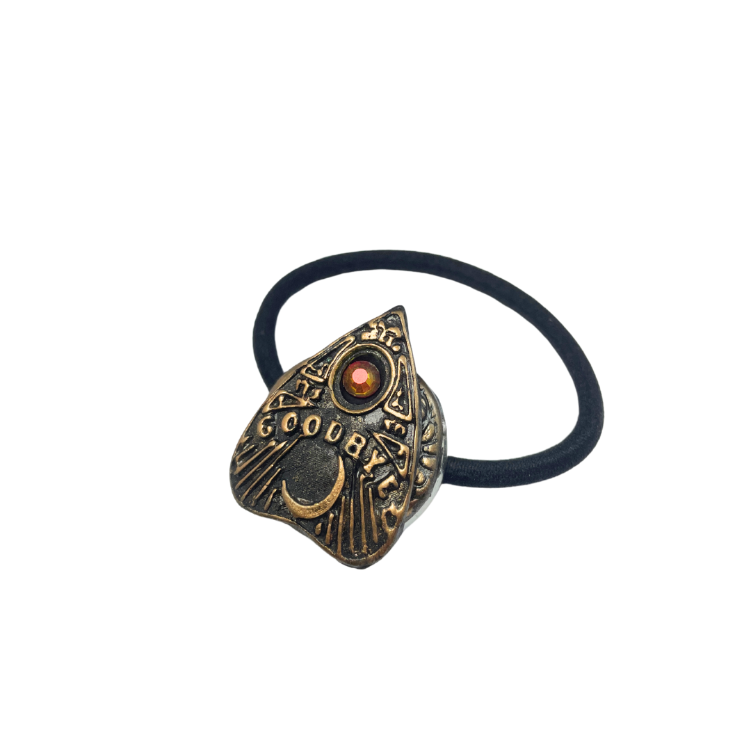Ouija board planchette hair tie with red crystal and the word 'goodbye' in gold lettering on a white background
