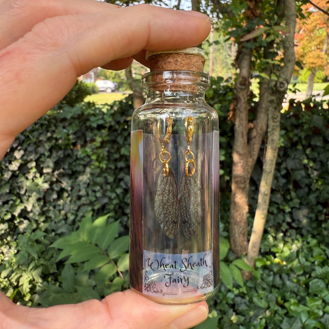 Hand holding a small glass bottle with cork lid containing fairy wing earrings, outdoors.
