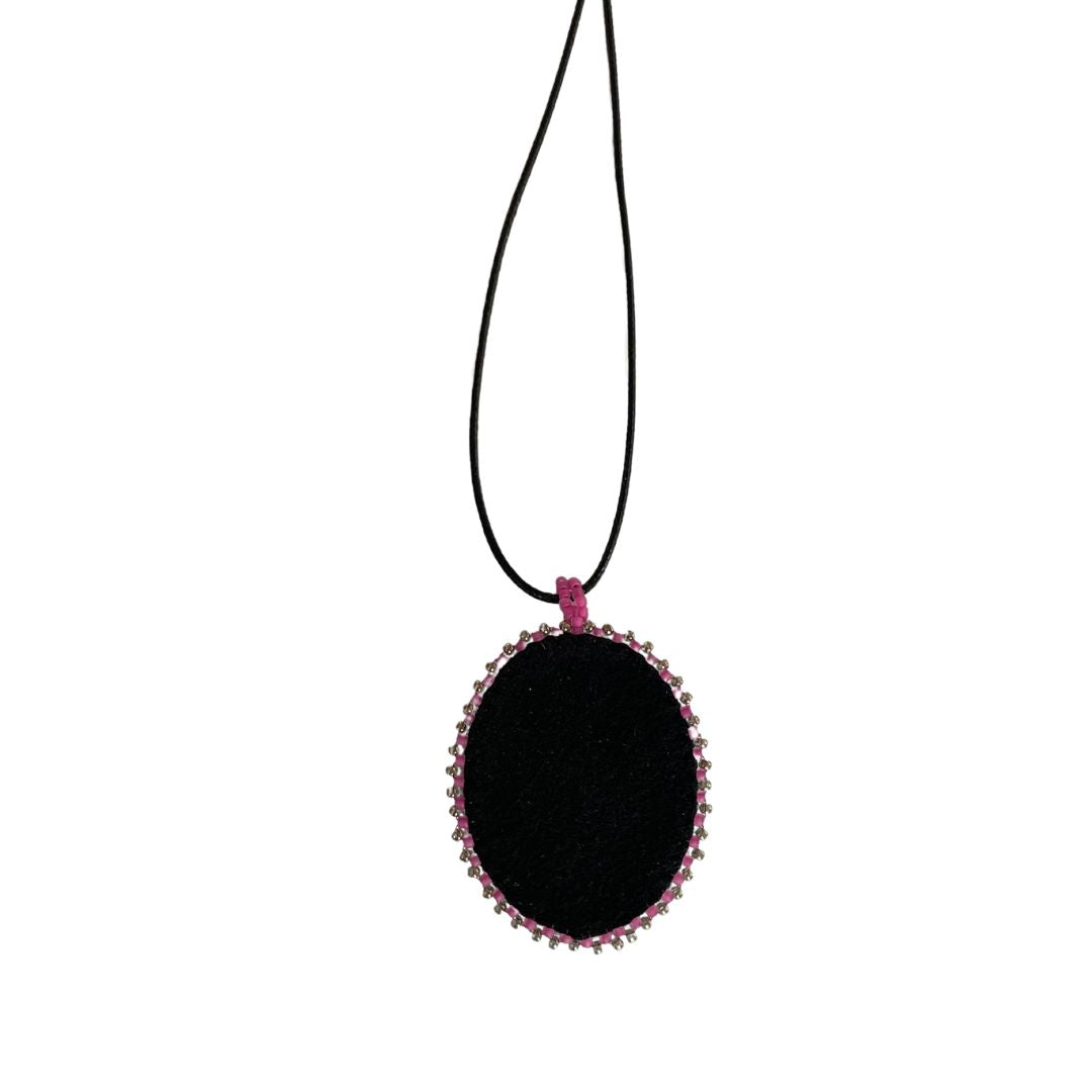 Black faux suede back of oval pendant with whimsical tree with purple, red and pink background with pink and purple glass seed bead edging on a black cord necklace.