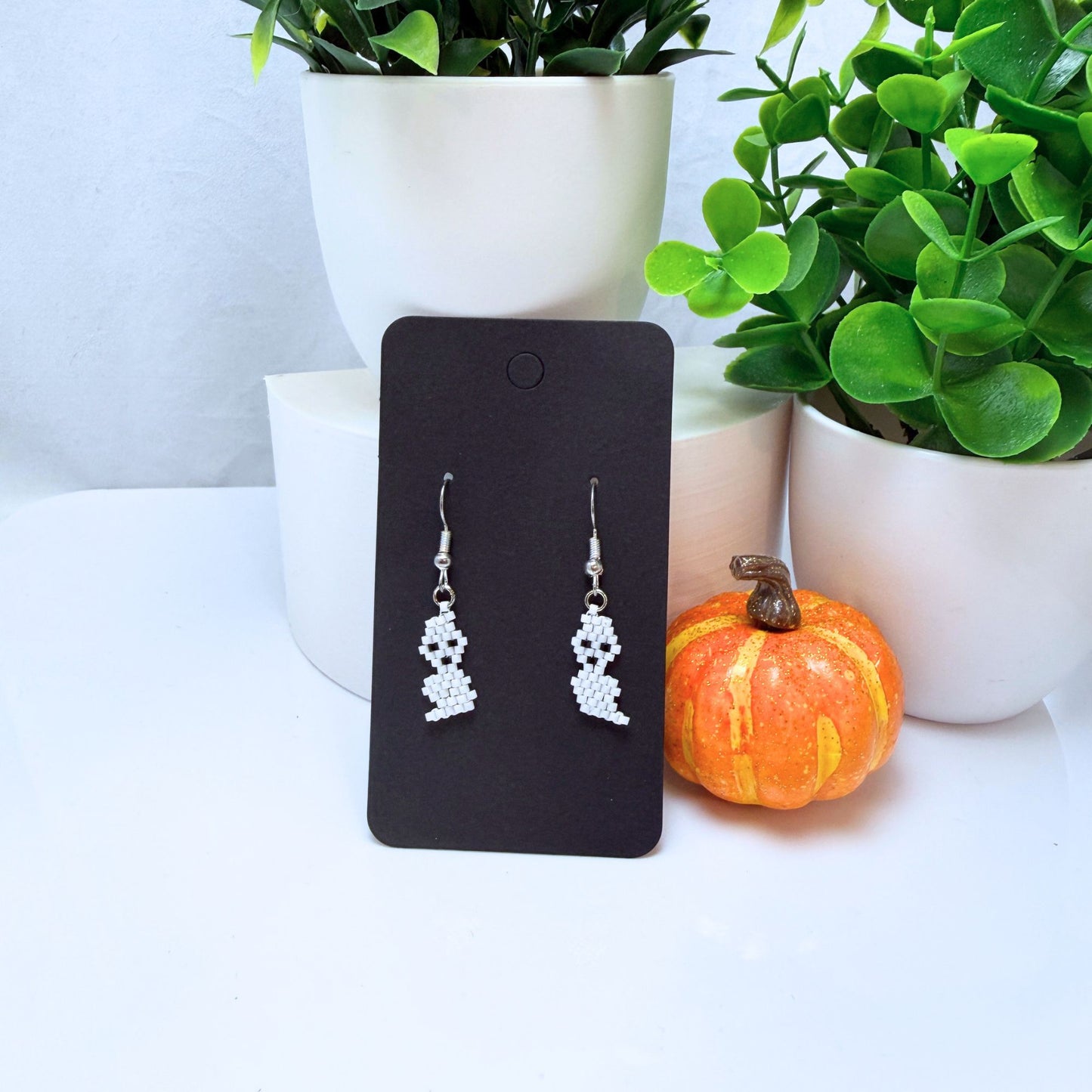 Beaded ghost earrings on black card displayed next to small orange pumpkin decoration