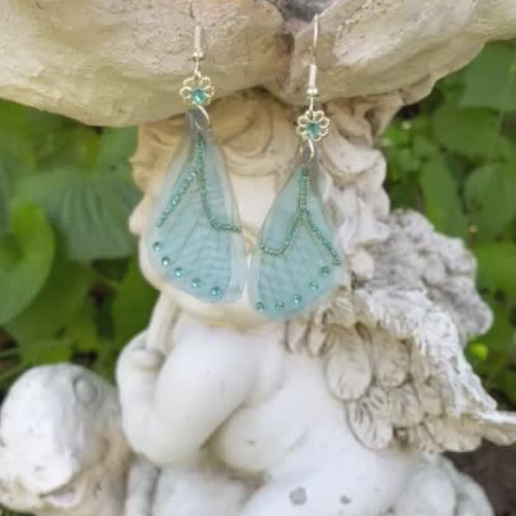 Blue chiffon butterfly wing earrings with silver crystal flower, charm and crystals on the wings hanging from a garden ornament.