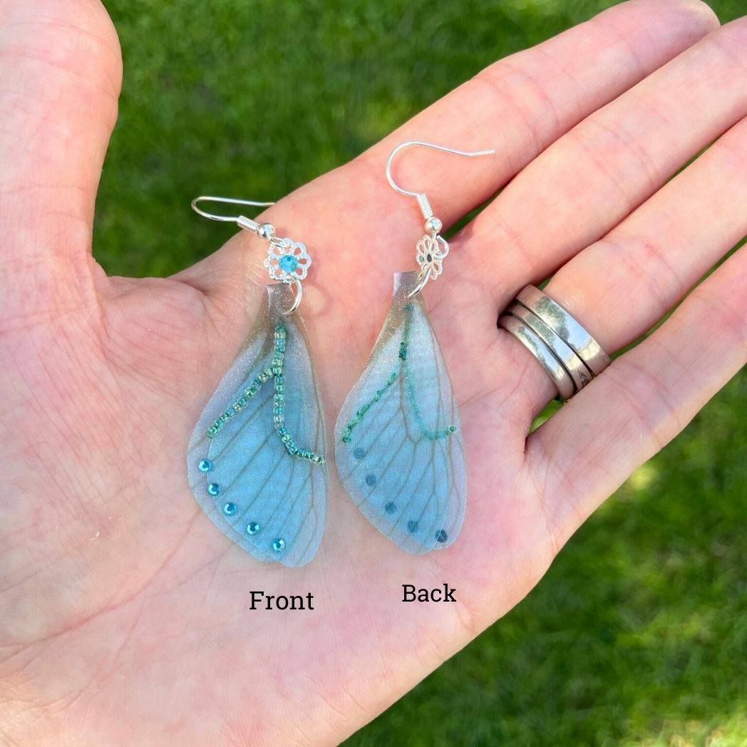 Blue chiffon butterfly wing earrings with silver crystal flower, charm and crystals on the wings hanging from a garden ornament. In a hand showing back a front of earrings.
