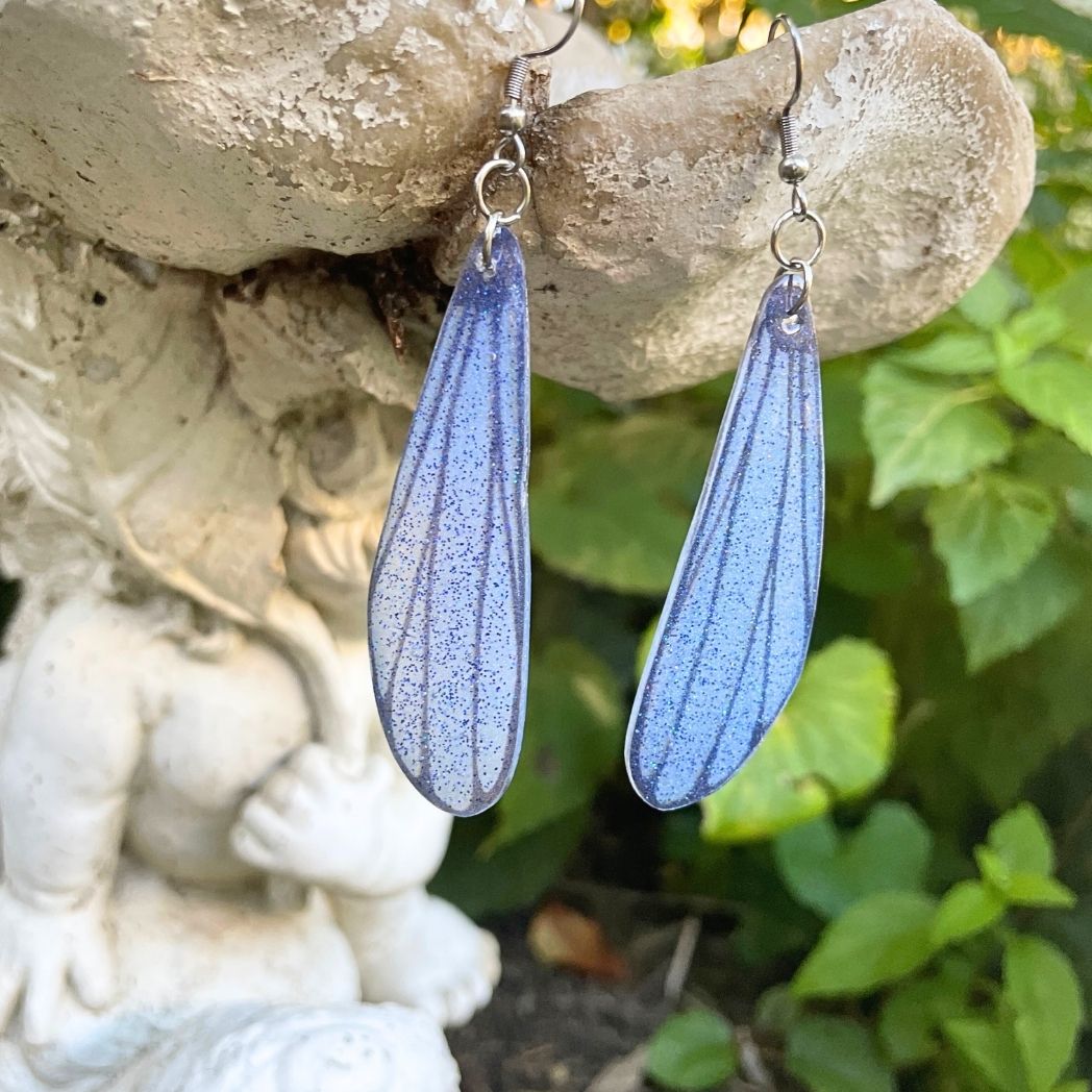 blue and gray sparkly long dangle dragonfly earrings dangling from a garden ornment.