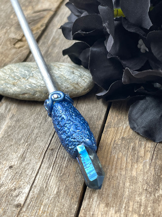 Blue raw crystal hair stick with hand painted clay details and crystals resting on a pebble with a black flower to the side.