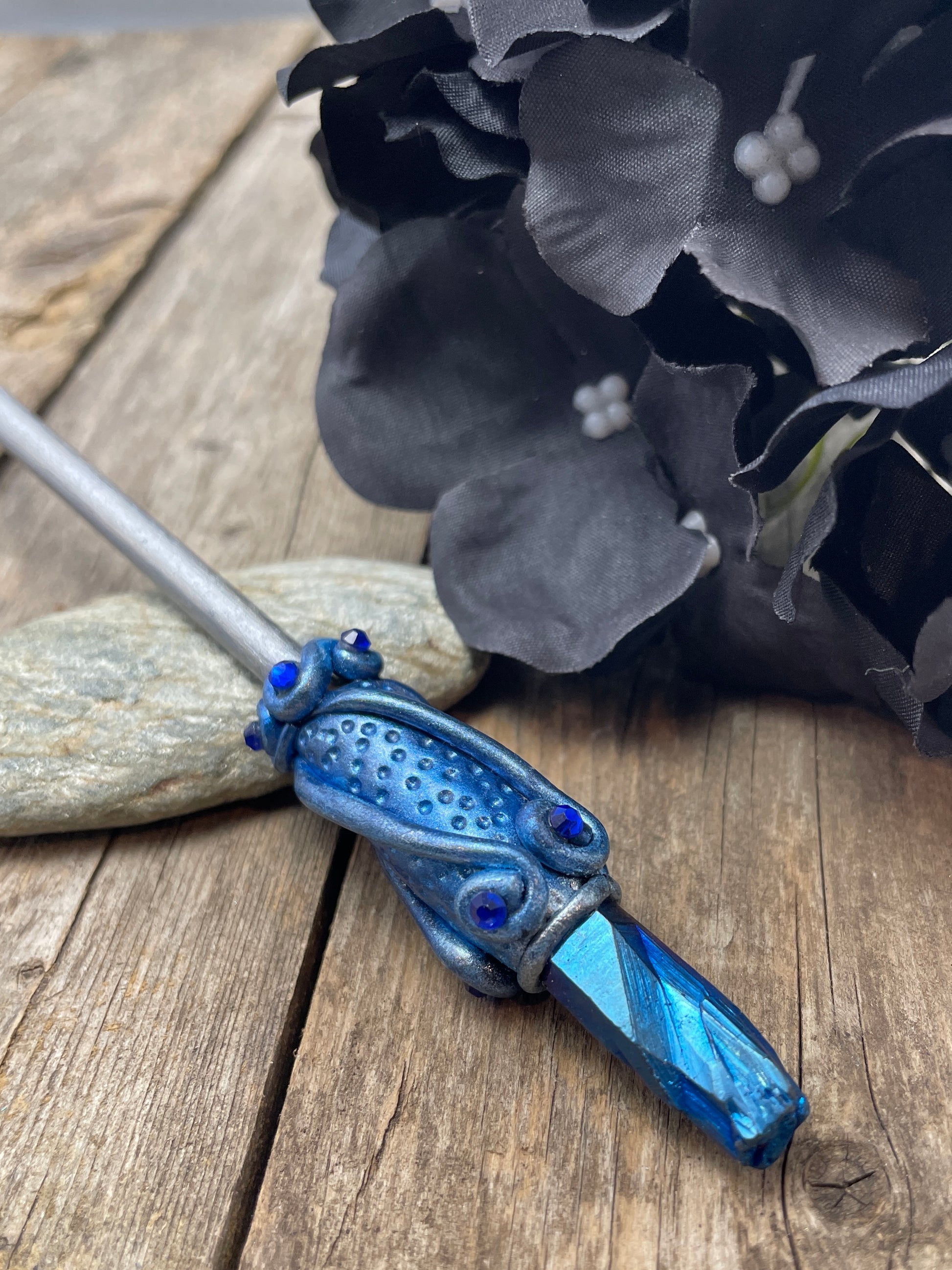 Blue raw crystal hair stick with hand painted clay details and crystals resting on a pebble with a black flower to the side.