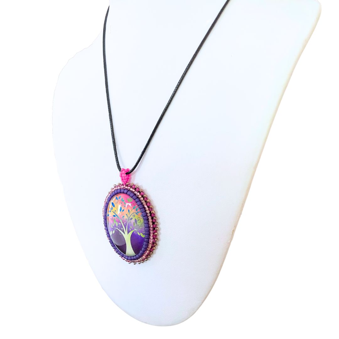 Oval pendant with whimsical tree with purple, red and pink background with pink and purple glass seed bead edging on a black cord necklace.