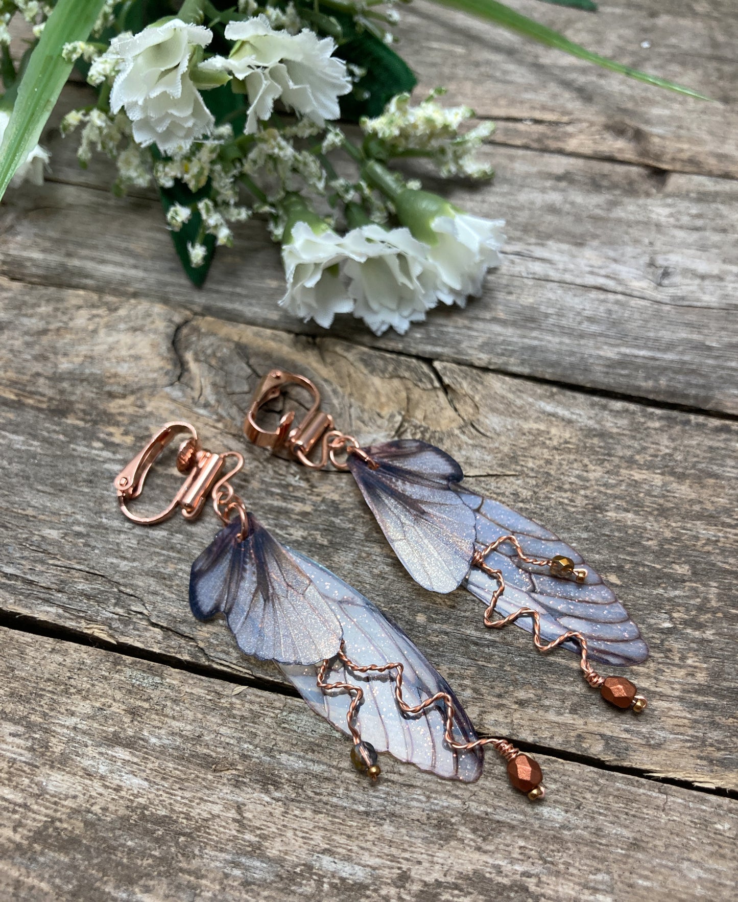 Fairy wing earrings with copper wired crystals with clip on components on wood surface with a flower next to them.