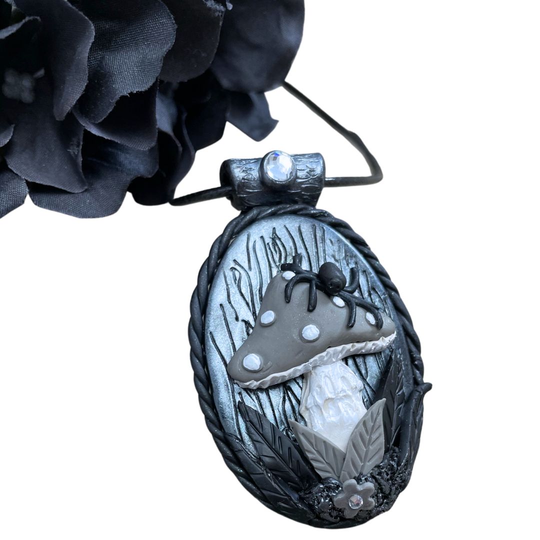oval pendant in silver and black with a spider sitting on a gray mushroom with flower detail at the base of the mushroom. A black flower is in the top left corner.