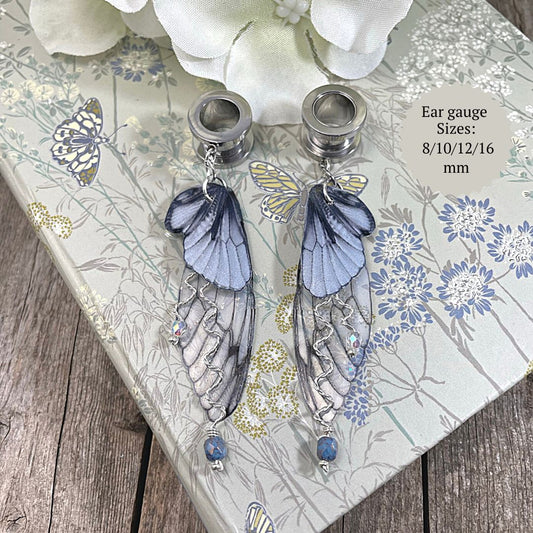 Double fairy wing earrings in silver with ear tunnels for stretched ears resting on a book with a flower in the background.