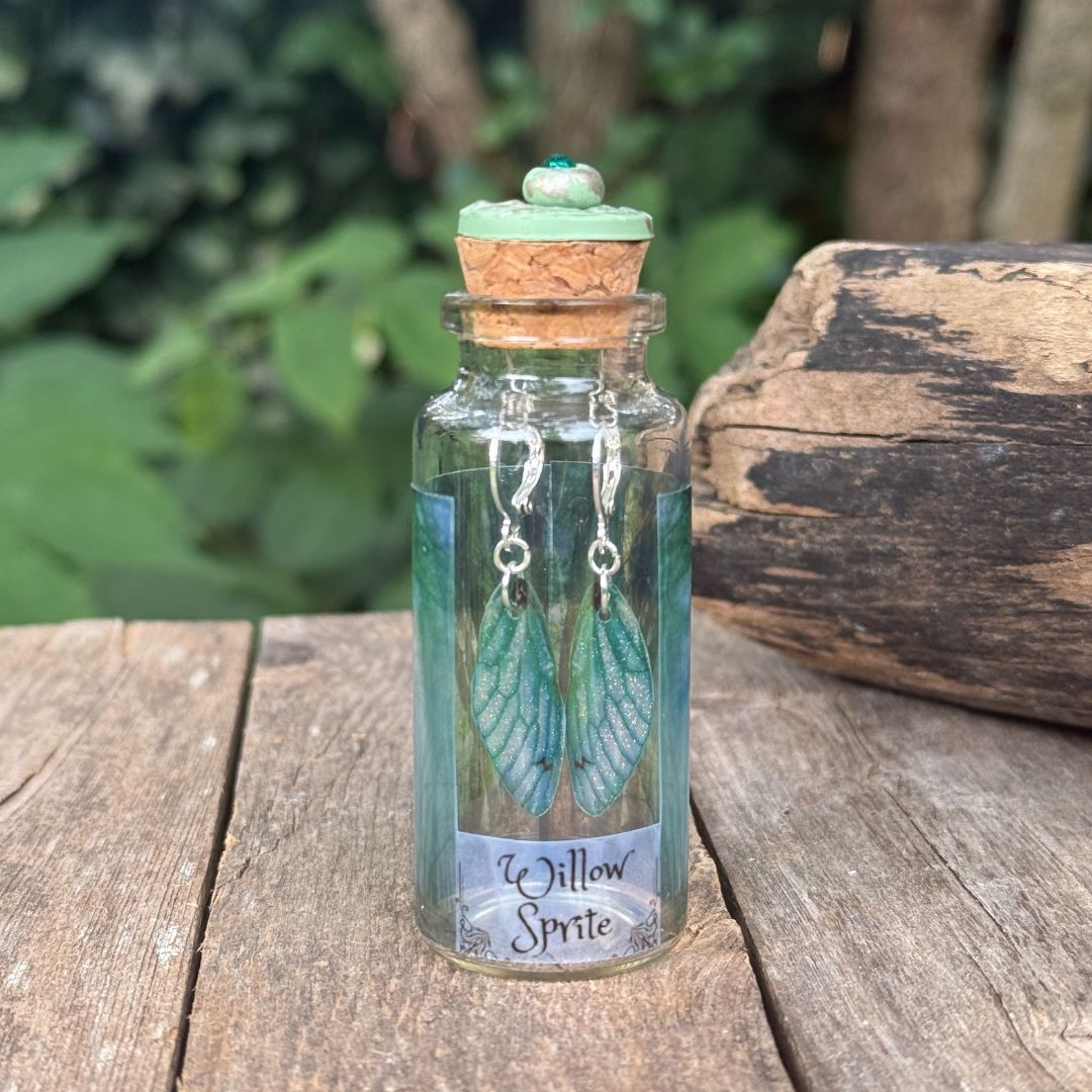 Glass bottle with cork lid containing green fairy wing earrings on a wooden surface with a natural background.