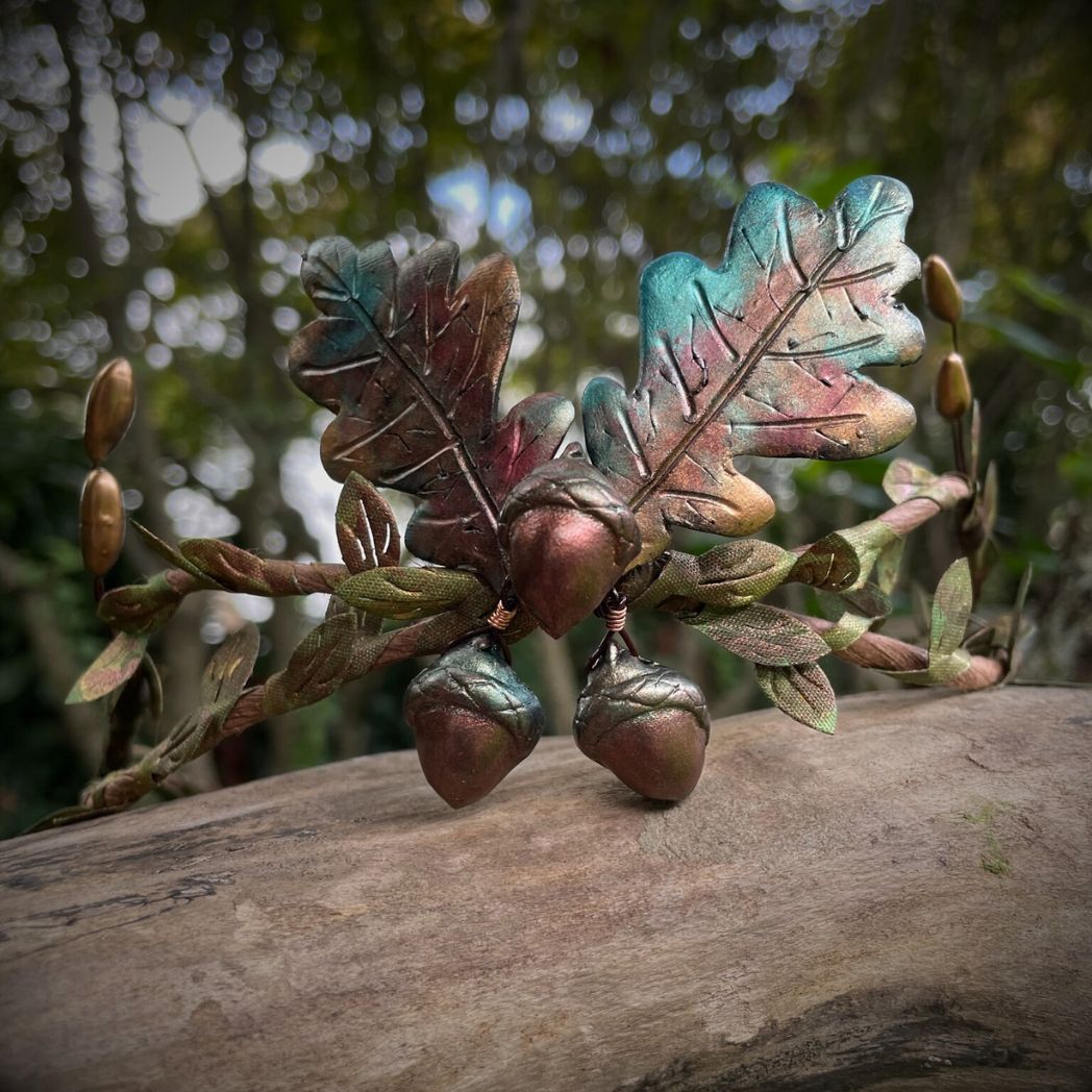 3 Acorns and 2 oak leaf crown in muted Fall colors with green and gold leaf vines and golden buds to the sides.  The crown in resting on a tree branch.