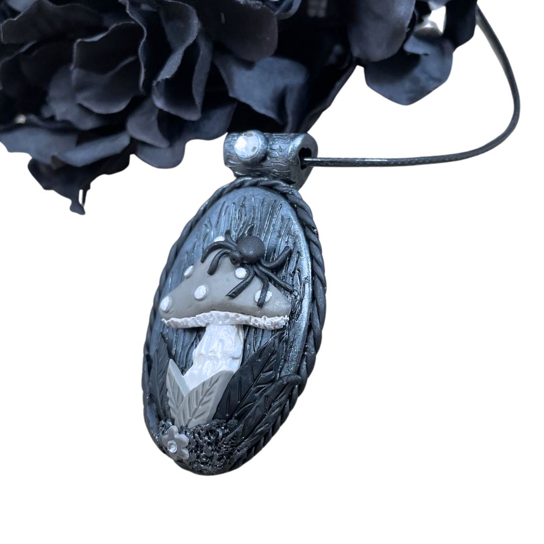 side profile of oval pendant in silver and black with a spider sitting on a gray mushroom with flower detail at the base of the mushroom. A black flower is in the top left corner.