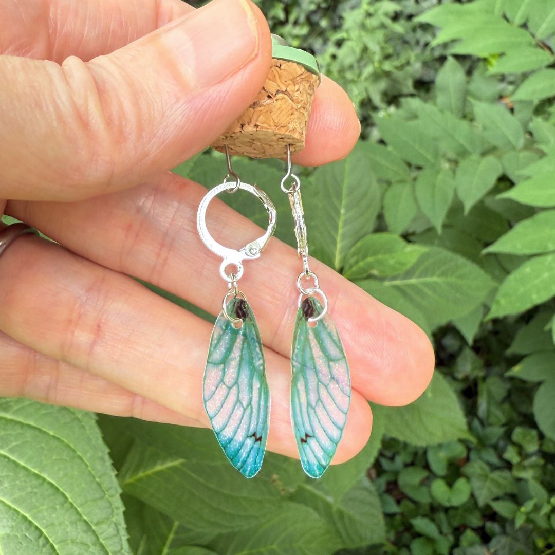 Green wing-shaped earrings held between fingers with a natural green leafy background