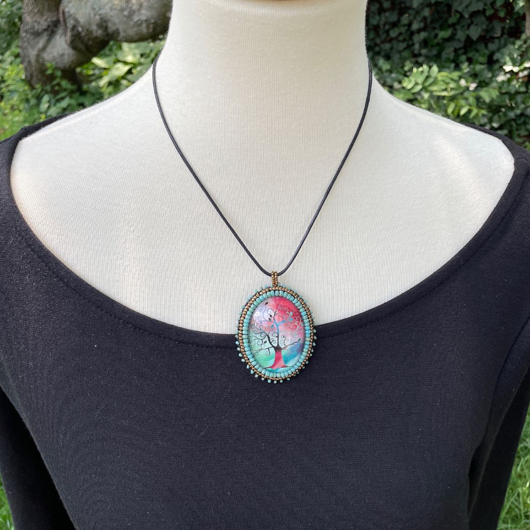 Silhoutte whimsical tree with red, blue and green background in an oval pendant with glass cabochon and green and gold seed bead edging on a mannequin,