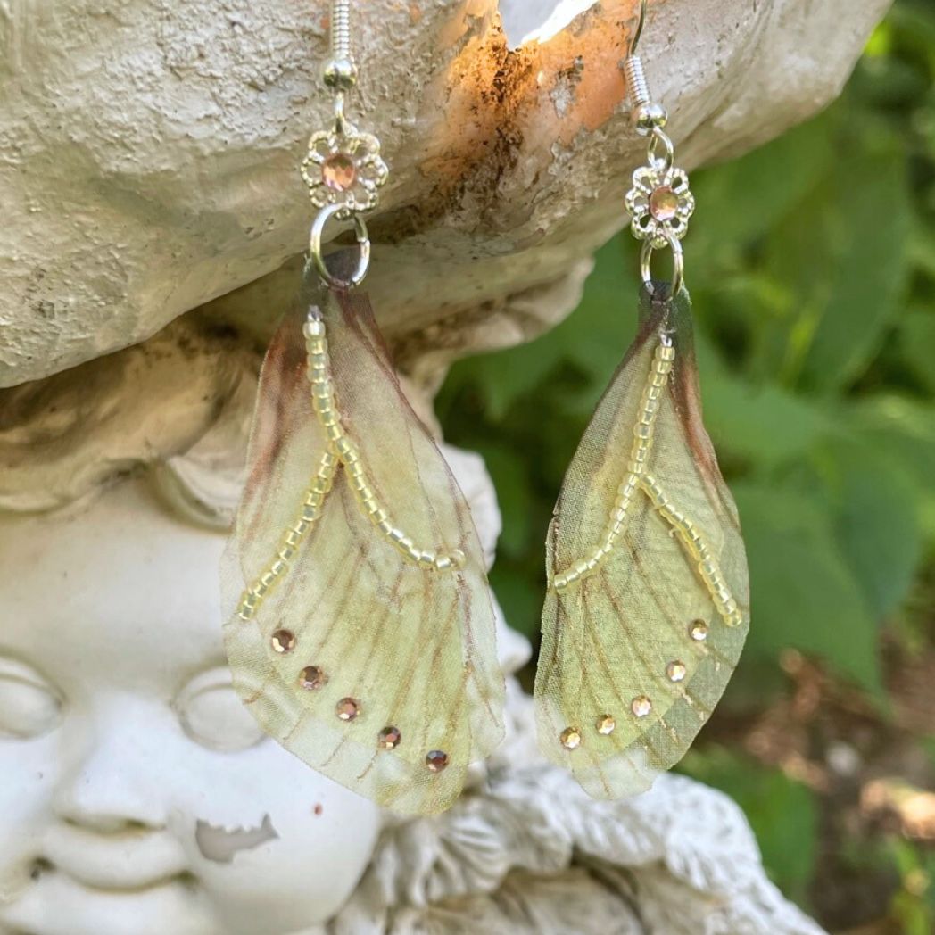 Yellow sheer fabric beaded butterfly wing earrings with peach color crystals and a flower bead connect the wings to the ear wires. Hanging from a garden ornament.