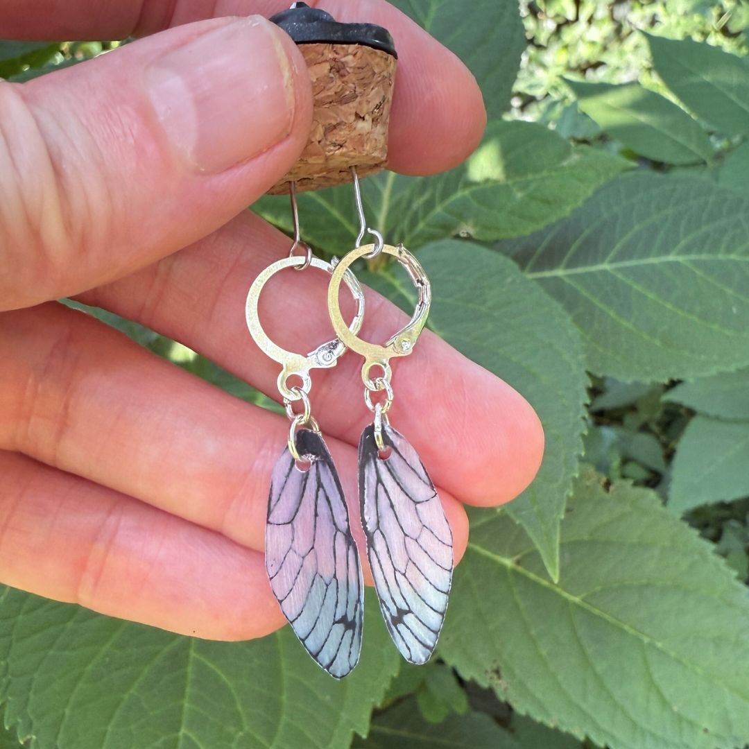Earrings with butterfly wing design held between fingers against a leafy background