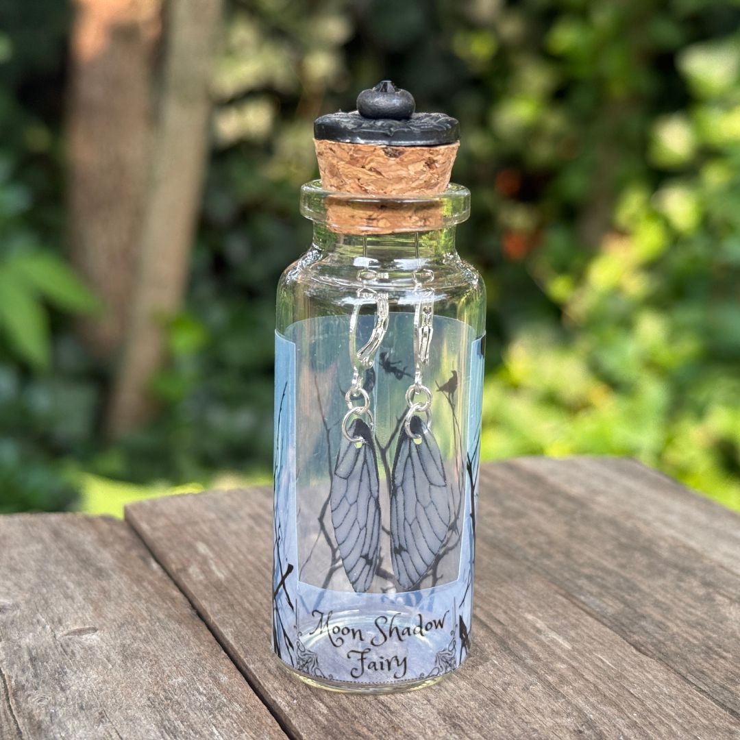Glass bottle with cork lid containing fairy wing earrings on a wooden surface with a blurred green background