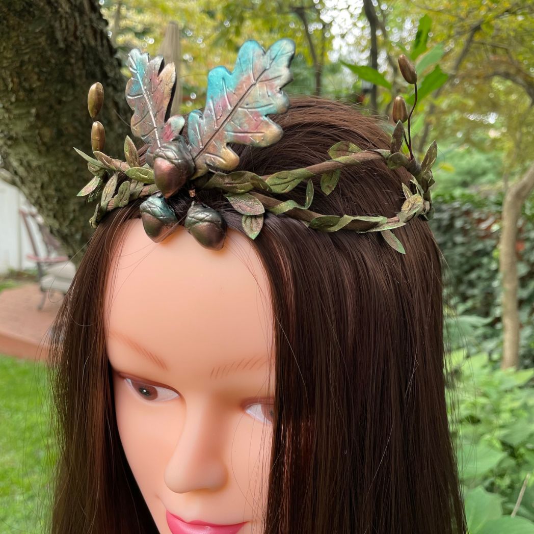 Left profile view of 3 Acorns and 2 oak leaf crown in muted Fall colors with green and gold leaf vines and golden buds to the sides.  The crown in displayed on a model