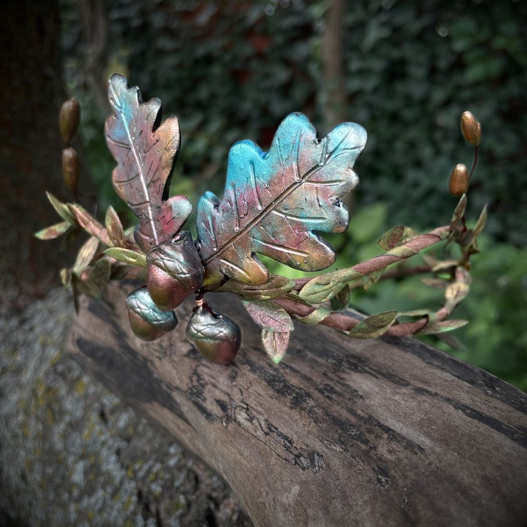 Side profile of 3 Acorns and 2 oak leaf  motif crown in muted Fall colors with green and gold leaf vines and golden buds to the sides.  The crown in resting on a tree branch.
