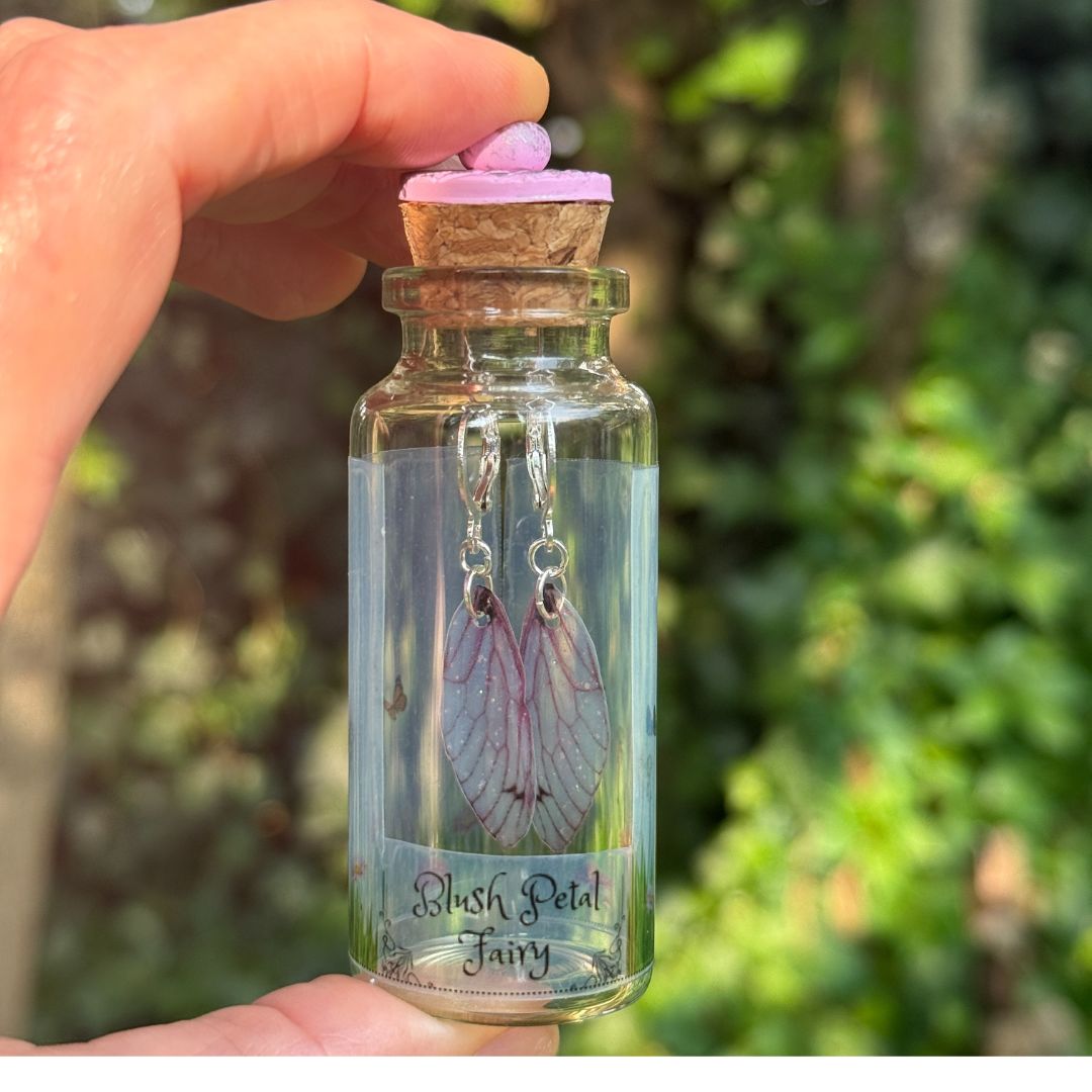 Glass bottle with cork lid containing 'Blush Petal Fairy' earrings, held by a hand against a blurred green background.