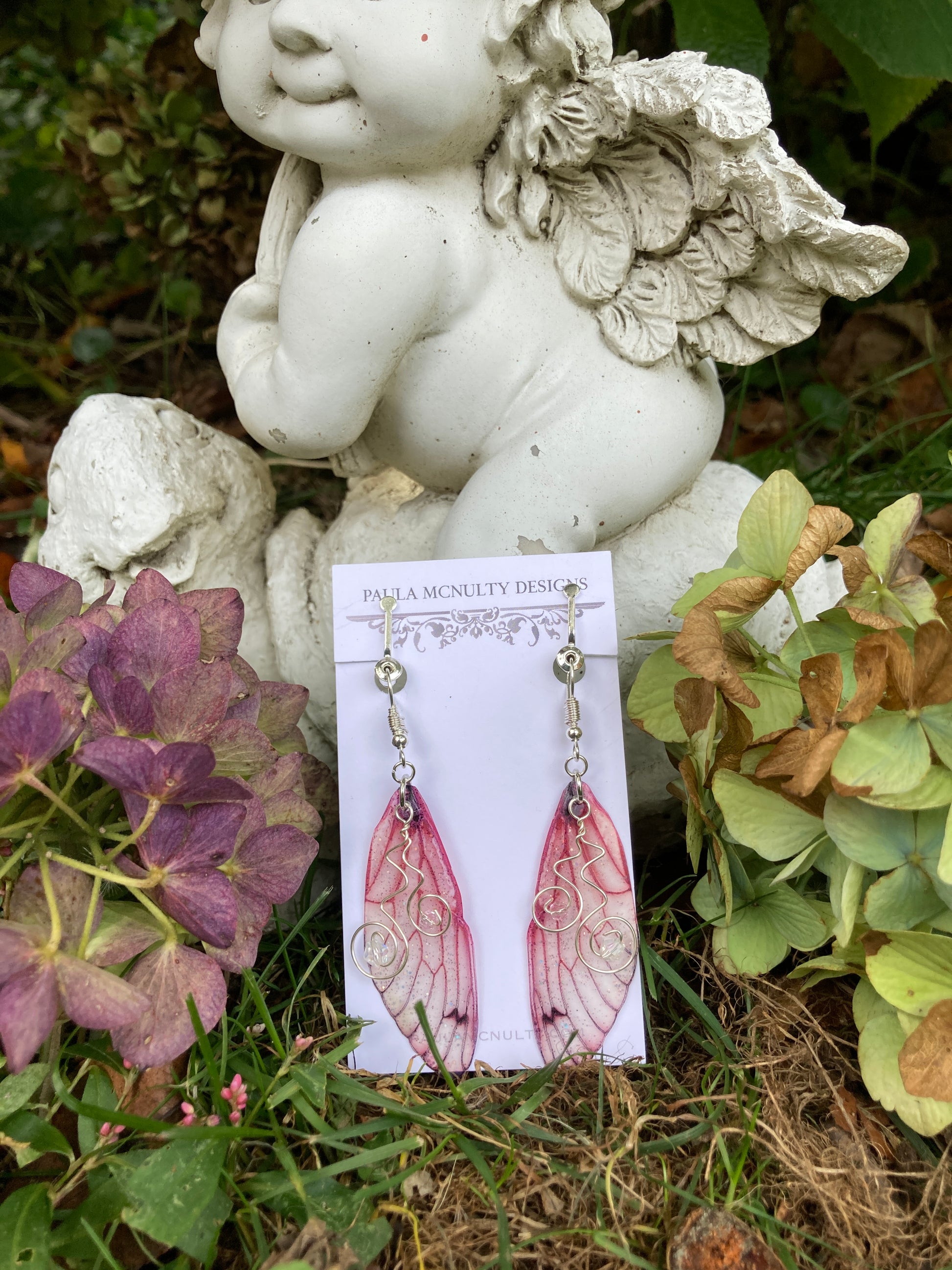 pink butterfly wing clip on earrings resting on an angel garden ornament with hydrangeas either side
