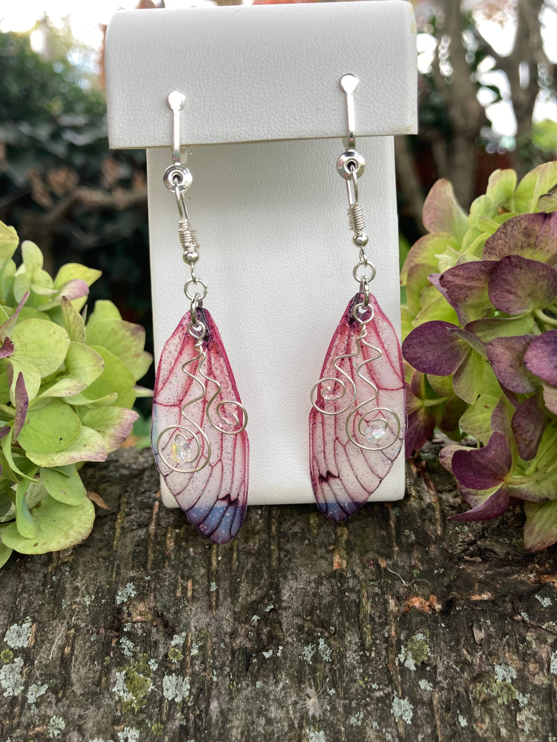 pink butterfly wing clip on earrings on an earring display outside with hydrangeas either side