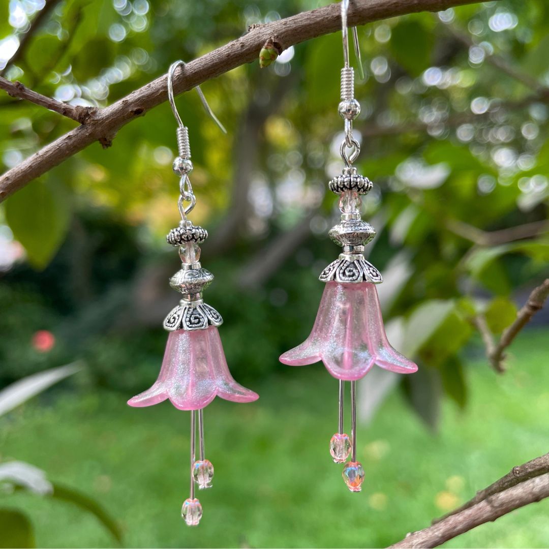 Pink Bell flower dangle earrings with pink crystal stamen with silver finish. Earrings are hanging from a tree branch.