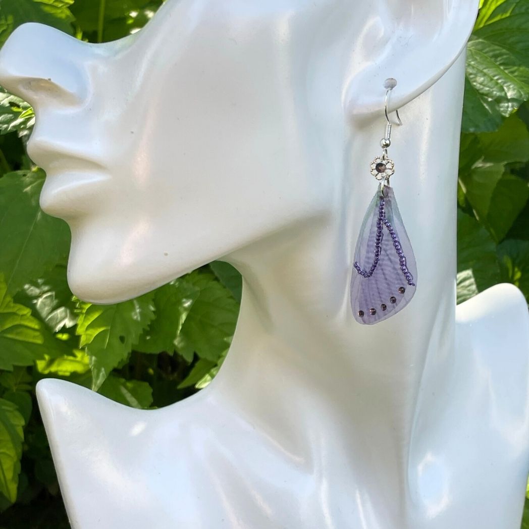 purple beaded fabric wing earrings with purple crystals and a flower bead with purple crystal above wings connecting to the ear wires on a model