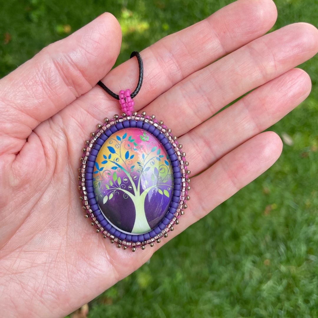 Oval pendant with whimsical tree with purple, red and pink background with pink and purple glass seed bead edging on a black cord necklace held in a hand