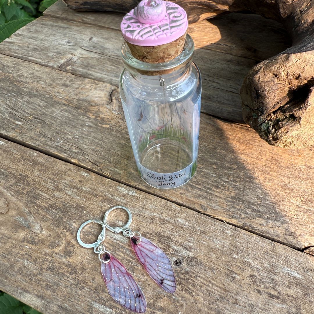 Clear glass bottle with cork lid on a wooden surface, featuring pink fairy wings earrings.