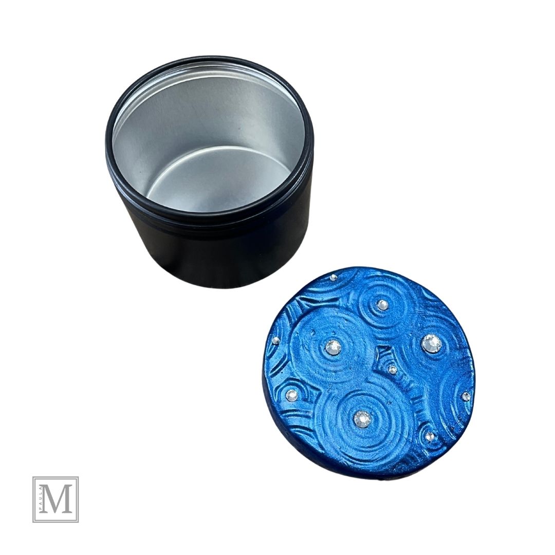 A black round tin with the lid to the side. Lid is decorated with polymer clay and hand painted metallic blue with blue and clear crystals.