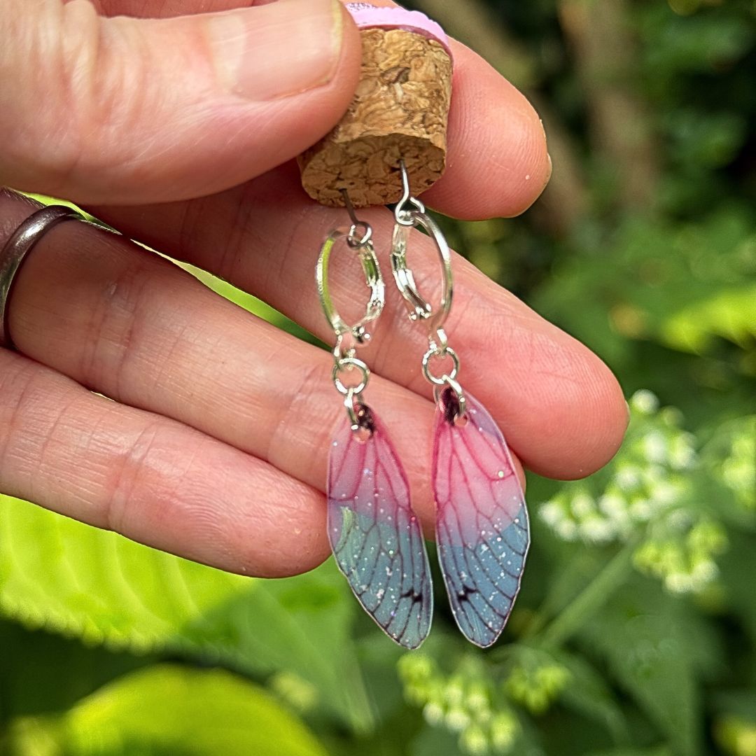 Hand holding a pair of pink and blue wing-shaped earrings with a natural background.