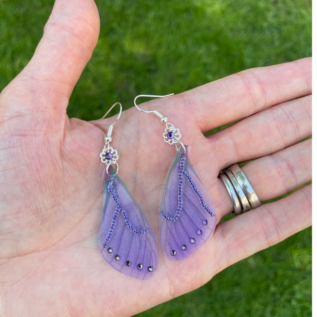 purple beaded fabric wing earrings with purple crystals and a flower bead with purple crystal above wings connecting to the ear wires held in a hand