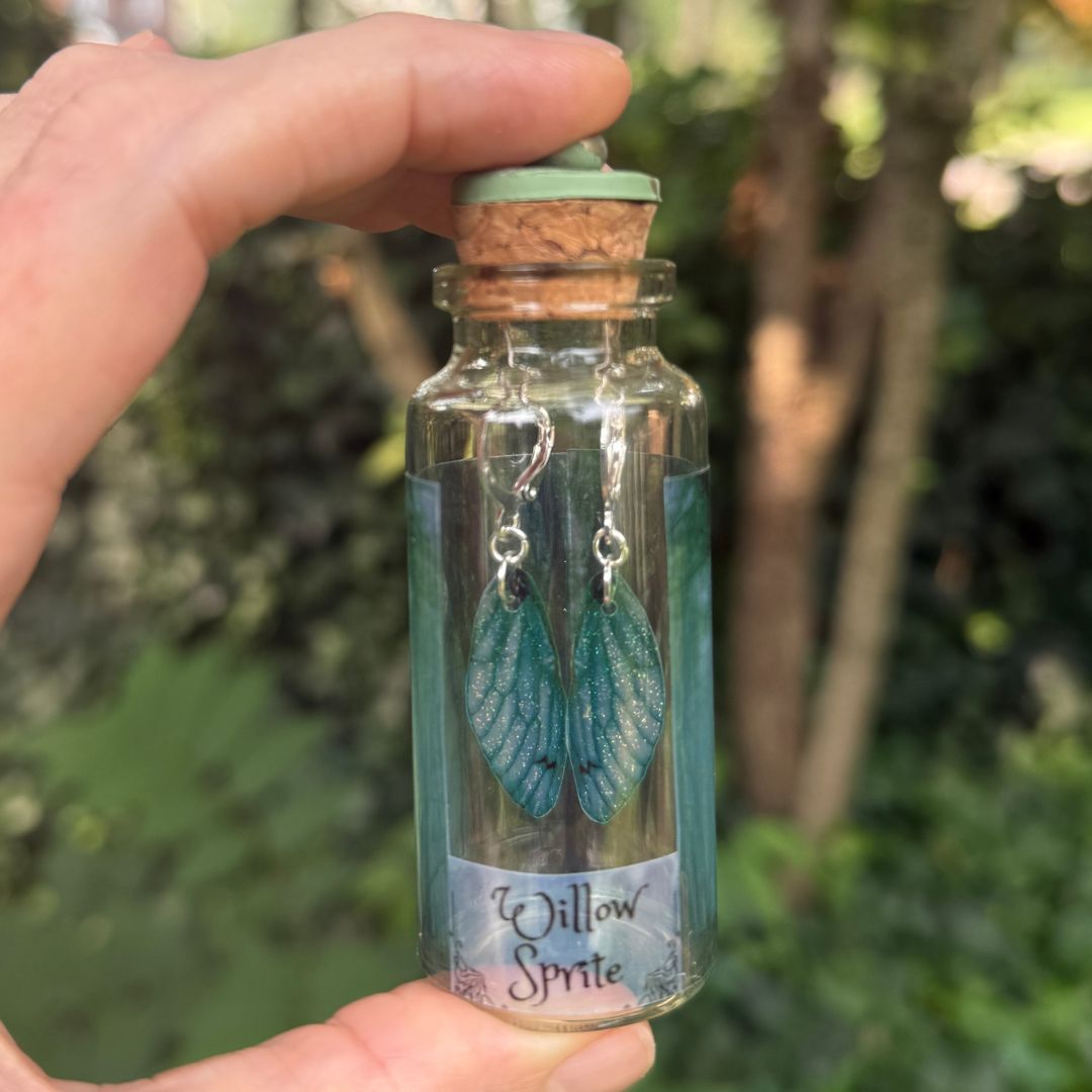 Hand holding a small glass bottle with 'Willow Sprite' earrings inside, against a natural background.