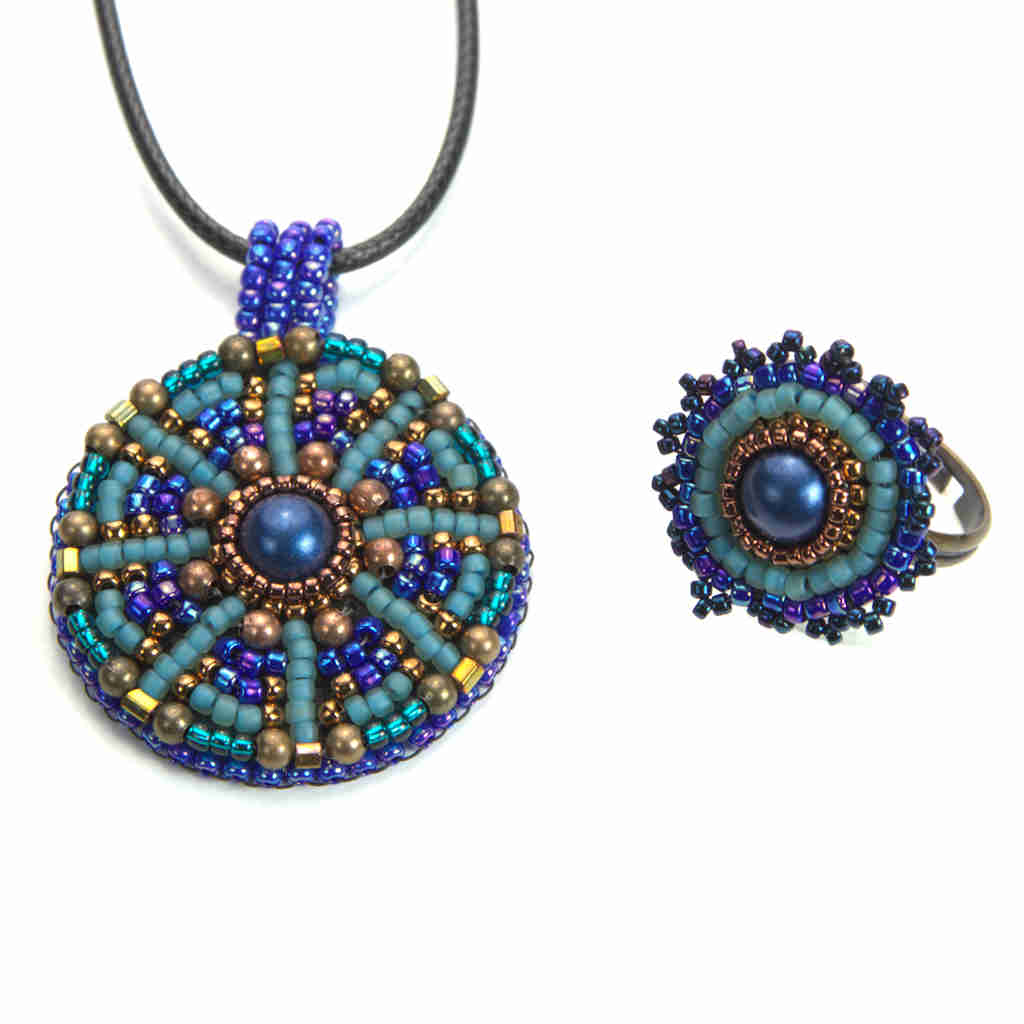 Close up of seed beaded mandala pendant necklace and ring set in blue and green resting on a pebble.