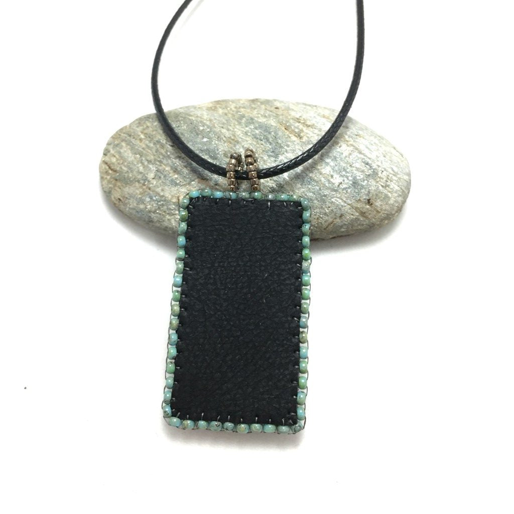 Back of patina faux leather floral textured rectangle pendant necklace resting on a pebble.