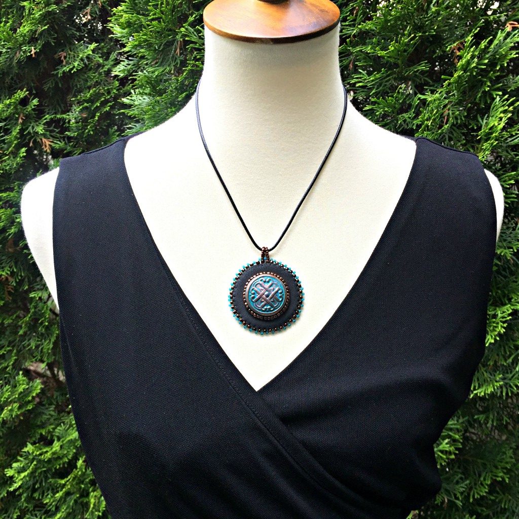 Model is wearing black faux leather round pendant with beaded edge in gold and turquoise with a patina Celtic knot motif in the center with beading. Necklace is on a black cord chain.