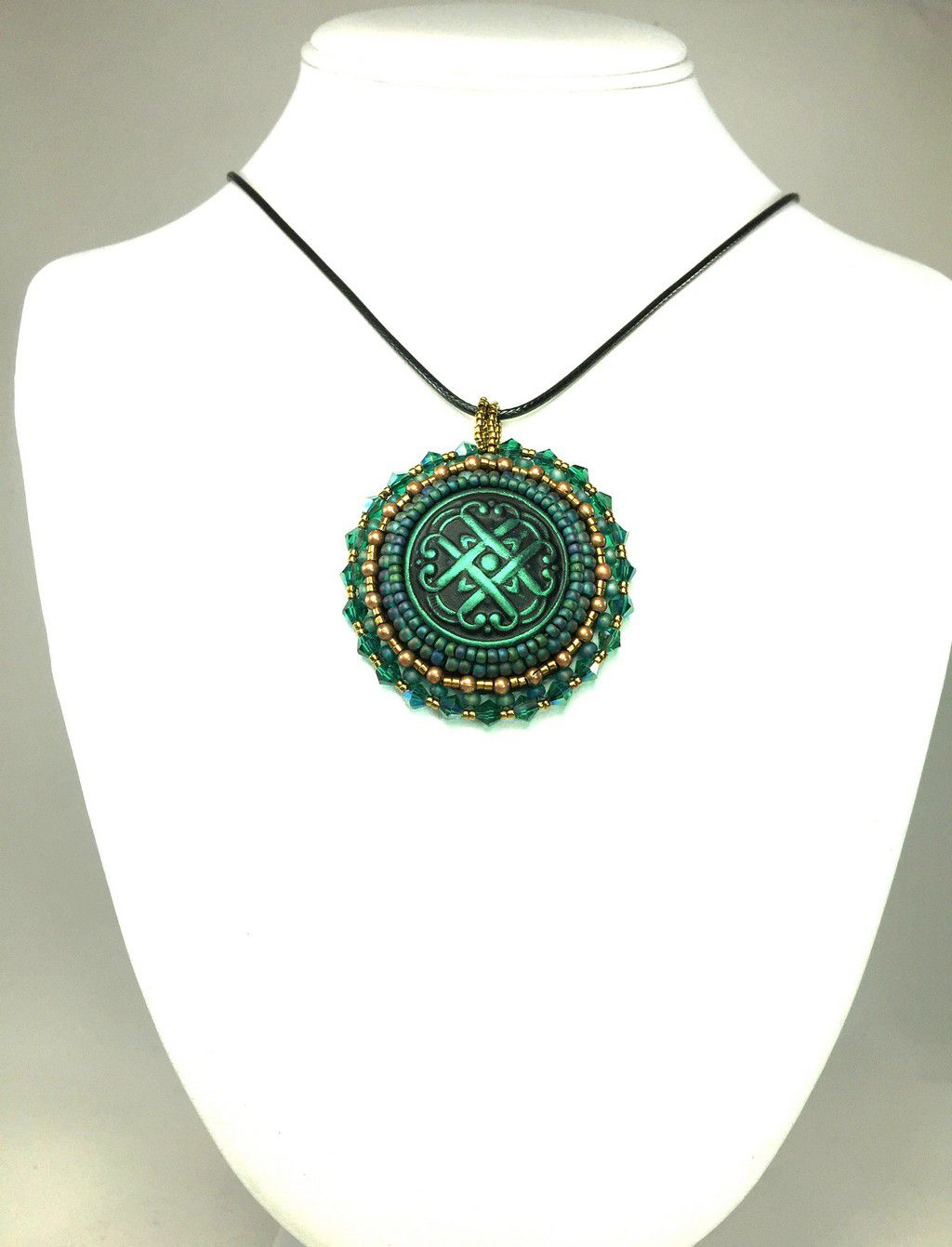 A green Celtic love knot motif, surrounded by green and gold seed bead edging  and green  crystals. Pendant is on a black cord chain on a display bust.