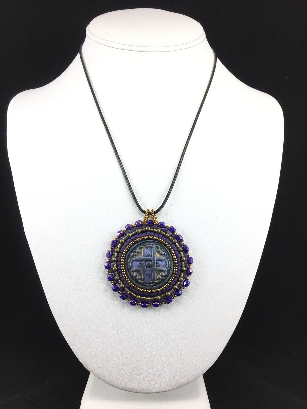 A purple Celtic love knot motif, surrounded by purple and gold seed bead edging  and purple crystals. Pendant is on a black cord chain on a display model.